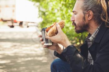 Bearded Asian man eating hot dog with coffee in summer park, outdoor profile portrait with selective focus