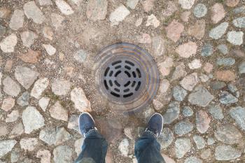 Male feet in dark blue jeans and sneakers standing on old street stone pavement in front of  sewer manhole