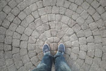 Male feet in dark blue jeans and sneakers standing on old gray stone pavement with round pattern