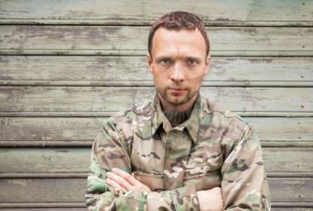 Young handsome Caucasian man in military camouflage uniform. Outdoor closeup portrait over green rural wooden wall