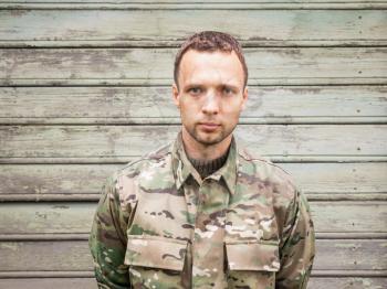 Young serious Caucasian man in military camouflage uniform. Outdoor closeup portrait over green rural wooden wall