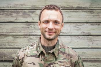 Young smiling Caucasian man in military camouflage uniform. Outdoor closeup portrait over green rural wooden wall