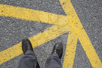 Male feet in leather shoes stand on on asphalt pavement with yellow road marking lines, first person view