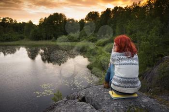 Teenage girl with red hair sitting on coastal stones near still lake in sunset