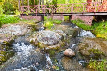 Small wooden bridge with red railings over stream with waterfall in summer park. Kotka, Finland