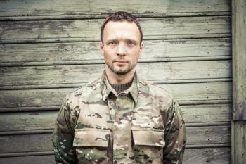 Young serious Caucasian military man in camouflage uniform. Frontal portrait over green wooden wall