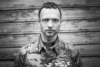 Young Caucasian military man in camouflage uniform. Closeup black and white frontal portrait over wooden wall