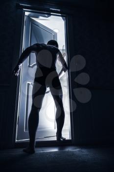 Young man enters opening door from black room and looks inside to the light, blue toned photo