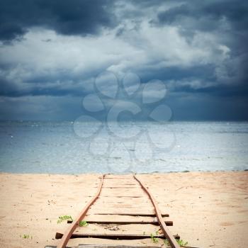 Old rusted railway goes over sandy beach to sea water under dark dramatic cloudy sky, useful for boat launching. Square photo with selective focus