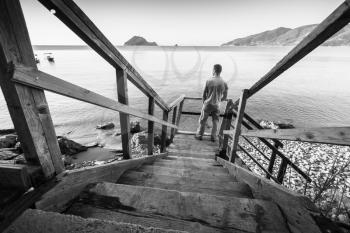 Young man stands on old wooden stairway going down to the sea coast in morning, black and white photo