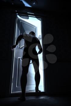Young man enters open door from black room and looks inside to the light, blue toned photo