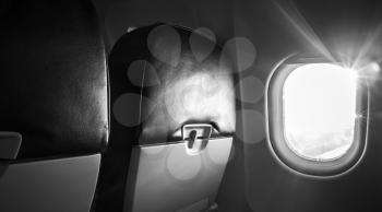 Passenger plane interior fragment. Main cabin chairs with folding table and glowing porthole with natural sunlight lens flare. Black and white photo