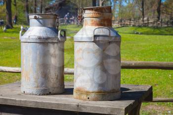 Two metal milk churns stand on wooden table over summer farm background