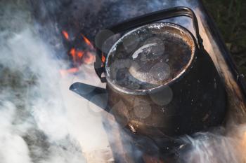 Old used black teapot with boiling water stands on a bonfire