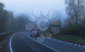Car with red stoplights goes on empty rural highway in dark autumn foggy morning, blue tonal correction filter effect. Shallow DOF with blurred background