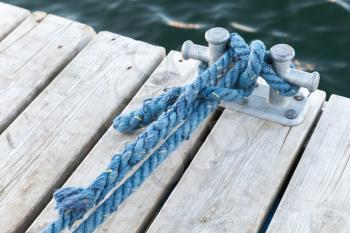 Mooring bollard with tied blue nautical rope mounted on white wooden pier, yacht marina safety equipment