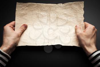 Strong sailor hands hold empty old crumpled paper sheet over black background, pirate map copy-space template