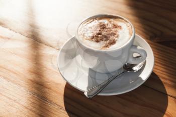 Cappuccino. Cup of coffee with milk foam and cinnamon stands on wooden table in morning sunlight