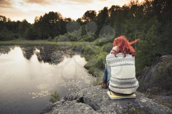 Teenage girl sitting on coastal stones near still lake in sunset, rear view