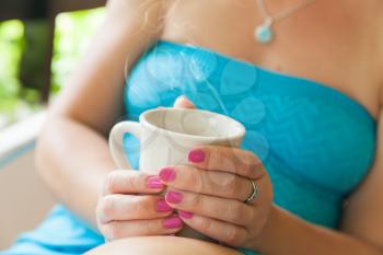 Young Caucasian woman in blue dress holds white cup of coffee in her hands. Close-up outdoor photo, selective focus on hands