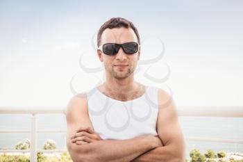 Young adult sporty Caucasian man in white shirt and black sunglasses stands with crossing hands. Outdoor summer portrait with sea on a background
