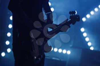 Live rock music background, electric bass guitar player in blue lights, closeup silhouette photo with soft selective focus