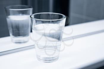Glass of water stand on white shelf near the mirror, blue toned close-up photo