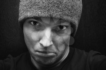 Young agressive Caucasian man in gray hat. Close-up studio face portrait over dark wooden wall background, selective focus, black and white photo
