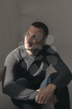Dark artistic studio portrait of young adult European man sitting in shadow under small sunlight beam over his face