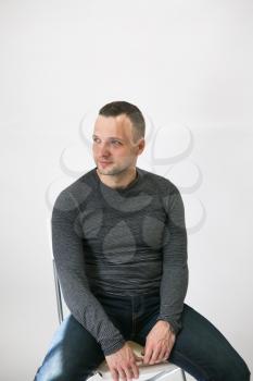 Young adult man is sitting on a chair near white wall, vertical studio portrait