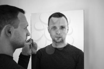 Black and white portrait of young adult European man standing near white wall with mirror