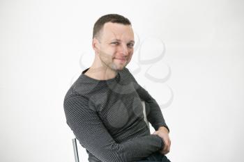 Young adult European man is sitting on a chair near white wall, close up studio portrait