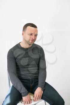 Young adult European man is sitting on a chair near white wall, vertical studio portrait