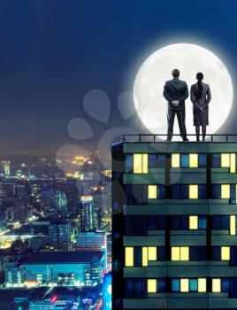 Man and woman look at the moon at night standing on the top of a high building
