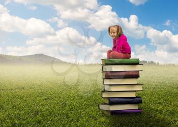 Small, cute girl sits on the pile of books on nature background