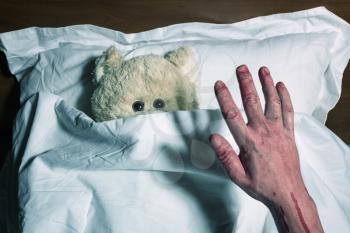 An adorable teddy bear laying in bed, scared by the bloody human hands, under the sheets.