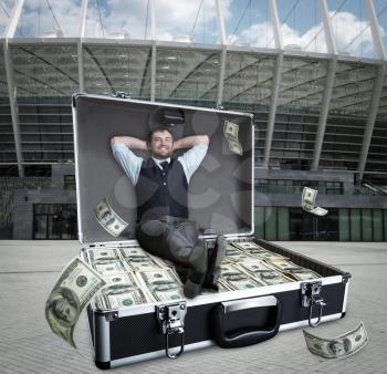 Happy businessman sits in the case full of dollars outdoors