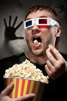 Portrait of scared man watching 3D movie and eating popcorn with dark hand behind
