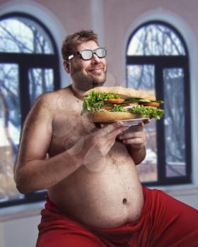 Fat happy man in glasses with sandwich sits in the room