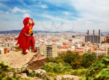 Little girl in a superhero costume standing on the rock against the city