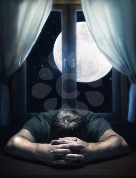 Sad man on the table against the moon in the window