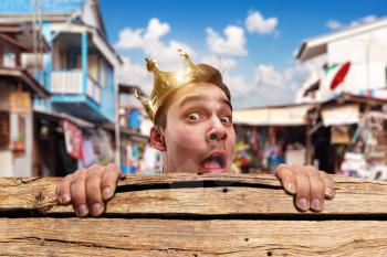 Scared pauper man with crown on the head hidding behind the wooden bench in slums