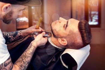 Young smiling man having his beard shaven, barber working with trimmer
