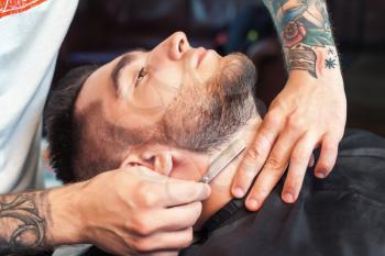 Young man having his beard shaven, barber working with razor in the salon