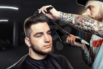 Young man having hair dress, barber working with hair dryer in dark room