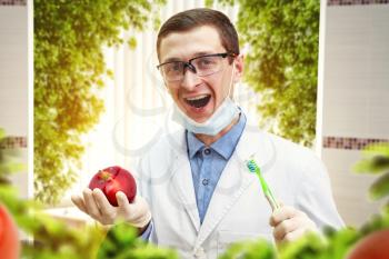 Young dentist holding tooth brush and an apple over green plants