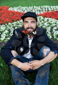 Bearded vagrant sitting in flowerbed with flowers in his beard
