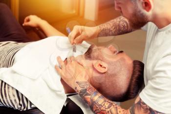 Young man having his beard shaven, barber working with razor in the salon