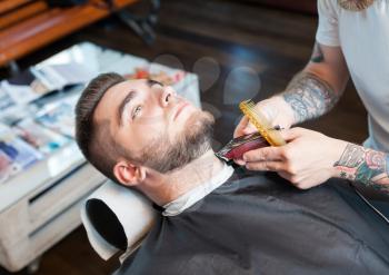 Young smiling man having his beard shaven, barber working with trimmer and comb