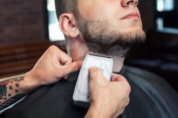 Young smiling man having his beard shaven, barber working with trimmer and comb